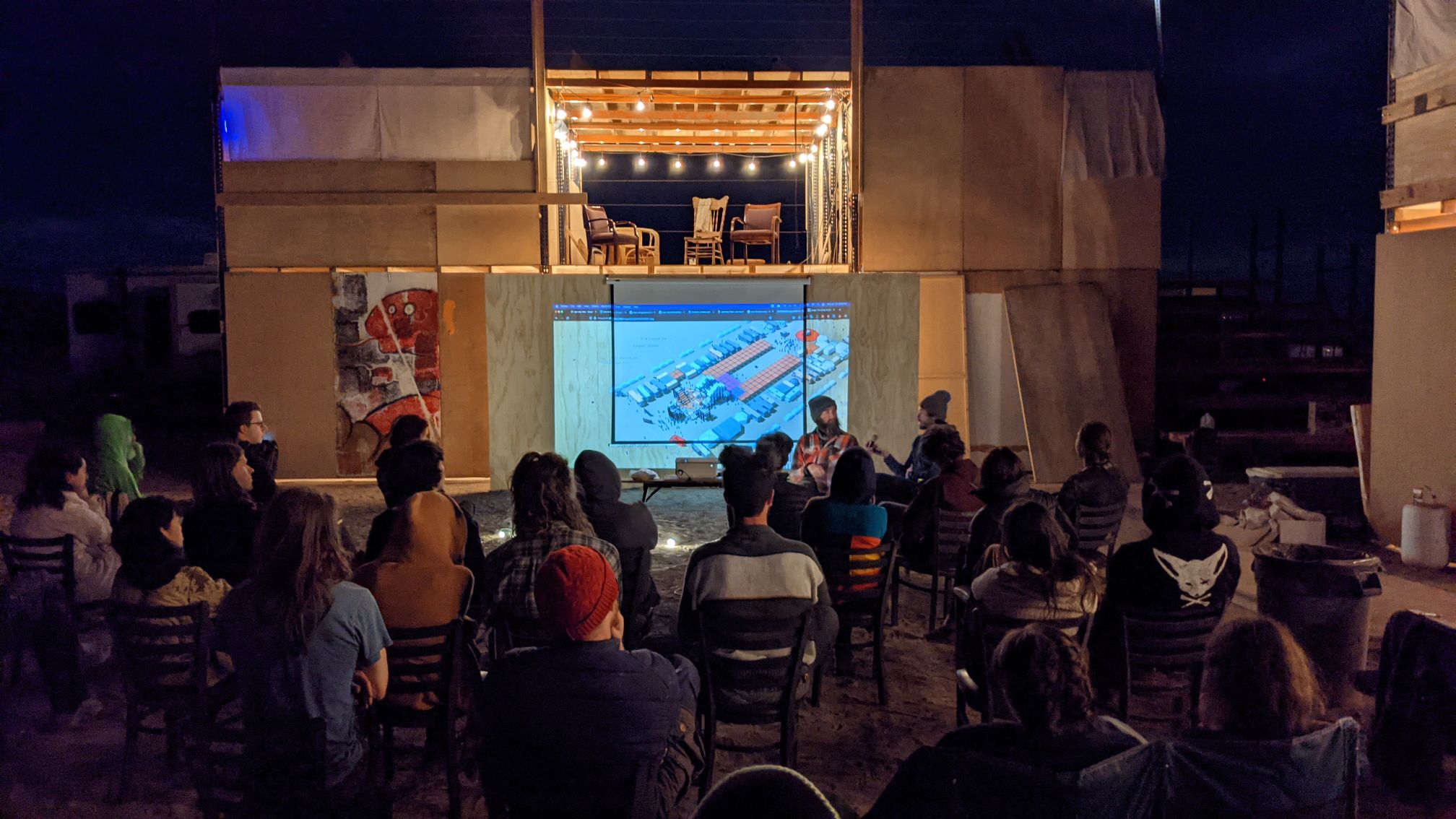 An audience watches a presentation at night in a self-built wooden structure.